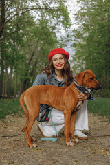 Portrait of cheerful woman petting brown Rhodesian Ridgeback dog in park and looking at camera 