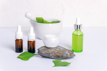 Fresh green ginkgo leaves, cream jar on a natural stone, brown glass bottles with essential oil, face serum bottle and mortar and pestle on white background. Modern apothecary, natural ingredients