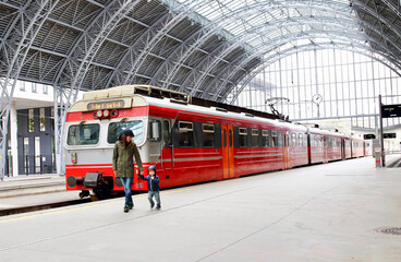 Young mother and little kid walk through the Bergen train station platforms with typical red Bergen-Voss line train on background. 