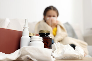 Girl blowing nose in tissue while on sofa in room, focus on medicines. Cold symptoms