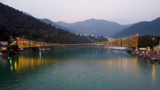 iron suspension bridge illuminated with ferry lights with ganges riverbank view at evening aerial video is taken at rishikesh uttrakhand india.