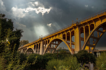 the Colorado Street Bridge surrounded by lush green trees and plants and powerful clouds at sunset...