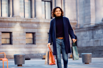 Young happy Hispanic man with shopping bags walking through city.