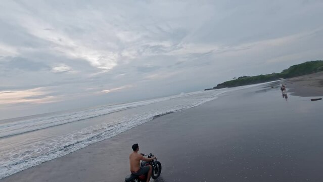 Asian man riding motorbike on black volcanic sand beach sunset tropical coastline enjoy freedom aerial view. FPV sports drone shot exotic shore sea landscape male speed driving motorcycle destination