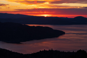 太陽光にオレンジ色の染まる空の色を弱く反射する湖。山と島の影。日本の北海道の美幌峠から見下ろす屈斜路湖。