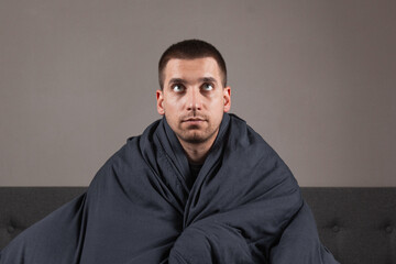 The image of a young man is sitting on a bed and covered with a dark blue blanket. He is holding a white cup of tea. The guy is looking at the camera. He is serious. Coronavirus and cold concept.