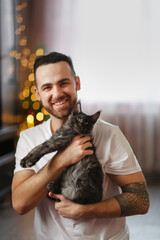 Obraz premium Portrait of manlike young man with beard with kitten in his arms. Kitten of Maine Coon kitten breed in arms of male owner. Christmas tree. Cozy Christmas time.