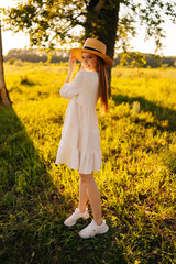 Vertical portrait of gorgeous redhead young woman in straw hat and white dress standing posing on beautiful meadow of green grass looking at camera, on background of warm sunlight at summer day.