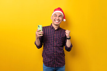 Excited young Asian man in Santa hat holding mobile phone, raising hands up, looking away at copy space over yellow studio background. Happy New Year 2023 celebration merry holiday concept