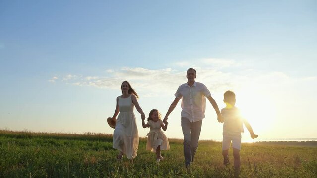 Happy parents with children run on green lawn at back sunset. Family enjoys walking together on meadow to watch sunset against blue sky
