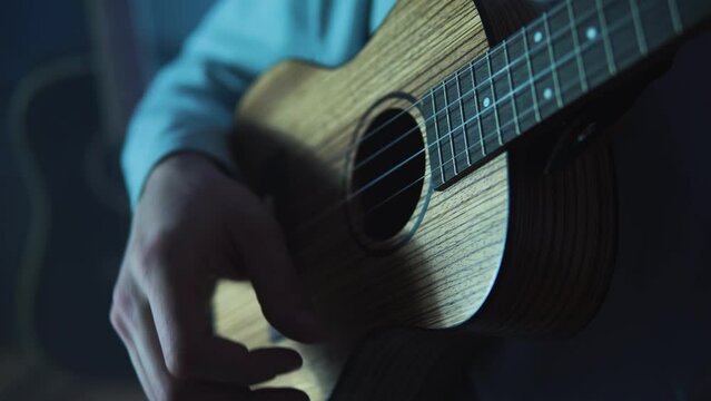 An adult smiling man plays the ukulele in the light neon dark room. A strong and blond guy sits and plays a small guitar in atmosphere place. A happy person plays a musical instrument.