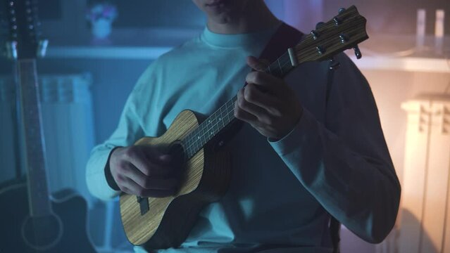 An adult smiling man plays the ukulele in the light neon dark room. A strong and blond guy sits and plays a small guitar in atmosphere place. A happy person plays a musical instrument.