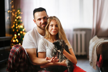 Cozy Christmas lovers. Cute traditional couple in pajamas with kitten on floor in living room by...