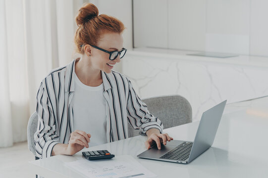 Young positive red-haired woman accounter working remotely on laptop and using calculator