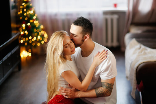 Young man tender kiss woman hugging on living room floor. couple in their red pajamas in living room by Christmas tree. Cozy good Christmas morning. Woman with big lips and long hairs.