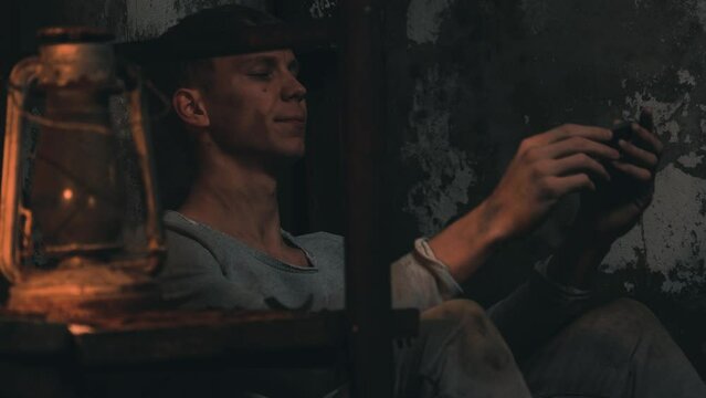 Tired and sad man deprived of liberty play on harmonica sitting in dark basement. blond guy wear dirty and torn clothes  Scary and old place in underground.