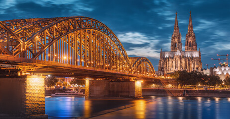 Panorama of illuminated bridge over Rhine river whith trains and tourists passing by and the Cologne Cathedral in the evening at the blue hour.