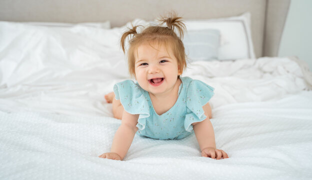 Happy Baby Girl Smiling In Blue Suit Pajama In White Bedroom Lying On Bed Looking At Camera. Cute Child With Pigtails. Childhood, Babyhood, People Concept. Copy Space Fro Advertisement