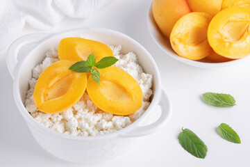 Fresh cottage cheese in white bowl with mint leaves and peach pieces and teaspoon on white background