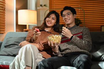 Happy young couple enjoying movies and popcorn, sitting on couch at home during New Year holidays