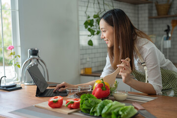 Asian housewife cutting fresh vegetables mixing salad ingredients cooking tasty vegan meal look search online blogger recipe notebook stand kitchen indoors
