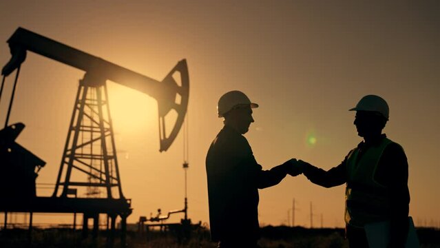 Teamwork.Oil engineer in helmet work with an oil pump.Silhouette of worker in field.Oil pump rig work at sunset.Two businessmen sign contract with handshake.Oil business concept.Work mineral deposit