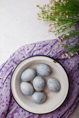 Blue painted Easter eggs in white bowl on purple kitchen table with flowers. Happy Easter concept. Vertical shot, top view, copy space