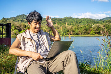 Young Asian traveling man relax and enjoy outdoor lifestyle camping in forest mountain on summer holiday. Attractive male relaxing in chair while talking calling on video on laptop