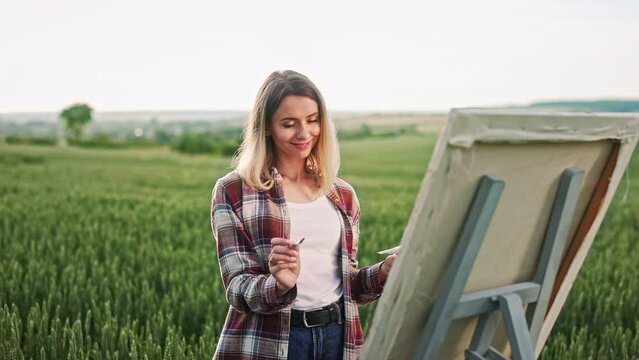 Side view of blonde woman wearing plaid shirt and jeans, painting outdoors. Female artist drawing on easel, holding brush and color palette, enjoying. Concept of art and painting.