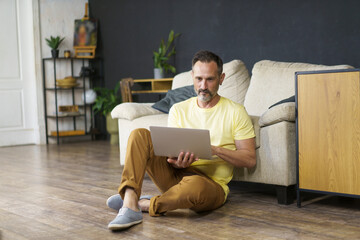 Fototapeta premium People and Technology Concept. Handsome mature man using laptop sitting on floor indoor lean on sofa watching video, movie or webinar, have a break