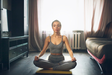 Pretty woman in gray sports top and leggings in lotus position meditates at home. Woman with long hair extensions and large silicone lips. Toning.