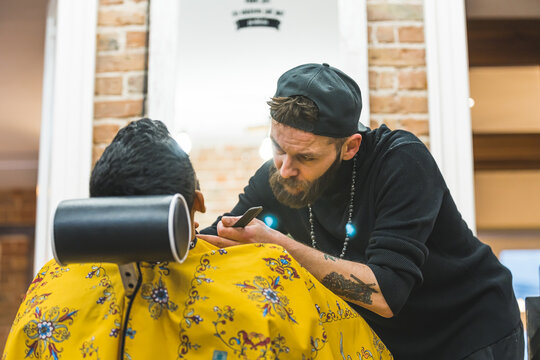 Back view of professional male barber with hat and tattoos trimming indian client's beard. . High quality photo