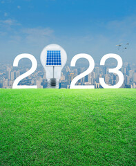 2023 white text and light bulb with solar cell inside on green grass field over modern city tower and skyscraper, Happy new year 2023 ecological cover concept