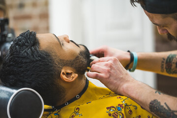 Professional barber with tattoos shaves the indian client's beard with comb and trimmer. High quality photo