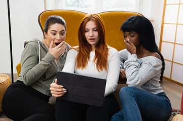 Multiethnic group of three young attractive women sitting in living room looking at digital tablet with surprised amazed face. Cheap prices of shopping online, test results at university or gossip.