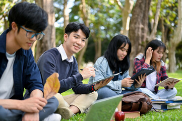 Handsome Asian male college student using his tablet while sitting in the university's park with his friends