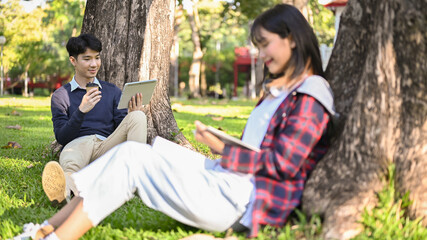 Handsome Asian male college student using his tablet and sipping coffee while relaxing in the park