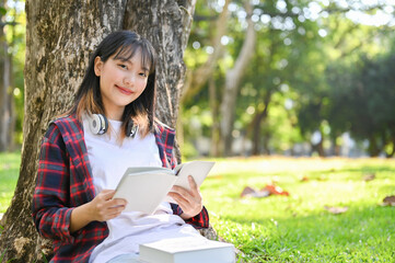 Beautiful Asian female holding a book, smiling and looking at camera while sitting under the tree