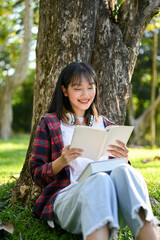 Portrait, Pretty Asian female reading a book while relaxes sitting under the tree in the park.