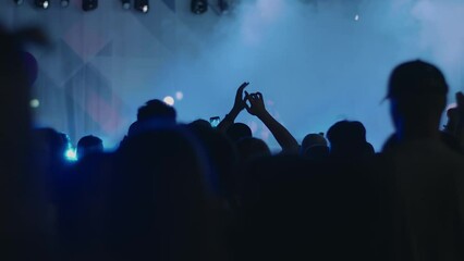 Silhouettes of crowd of people dancing with raised hands at concert in front of stage with blue smoke and spotlights. Back view - Powered by Adobe