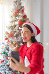 Cute beautiful young asian lady woman wearing santa hat selfie and video call with mobile phone posing in front a big Christmas tree with lots of decoration lights gift box and ornaments