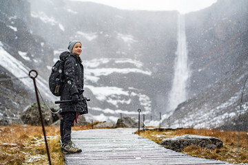 youn woman smiling, looking in camera and holding a camera in hand on stick on wooden trail at Hengifoss waterfall with layers of red clay during falling snow