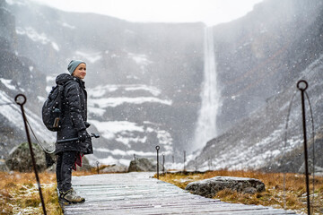 youn woman smiling, looking in camera and holding a camera in hand on stick on wooden trail at Hengifoss waterfall with layers of red clay during falling snow