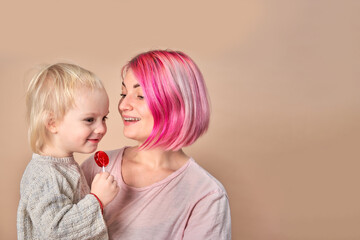 A young modern mom with pink hair holds a baby with a lollipop on a light background .Individuality, Motherhood, Parenthood, vacations.