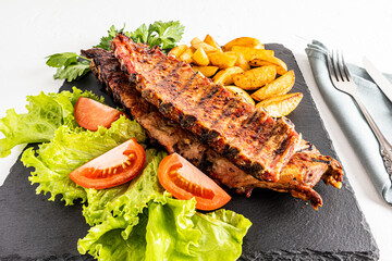 a large kind of pork ribs on a slate board with lettuce greens, slices of tomato and grilled potatoes. the concept of food cooked in the backyard.