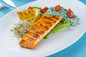Grilled salmon steak. A piece of red fish on a white plate and a blue background on the table. Fish food with sauce, tomatoes, lemon. Restaurant menu. Grill fried