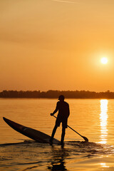 Fototapeta na wymiar Silhouette of a man paddle on stand up paddle board SUP against autumn orange sunrise on river