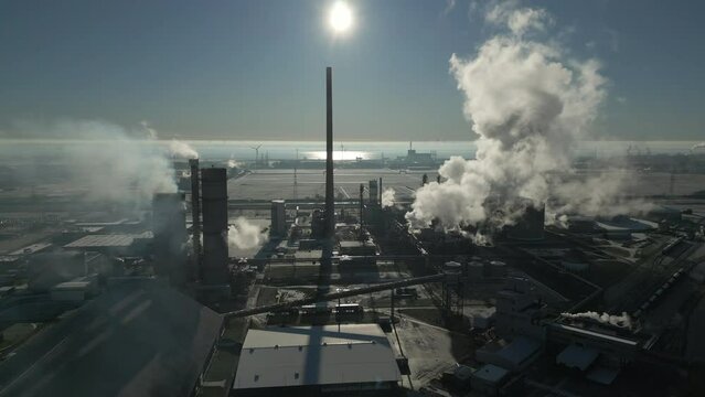 Smoke and steam rising of an industrial facility, CO2 emissions, air pollution, exhaust gases, aerial drone shot, backlit scene, sunny