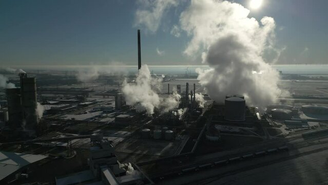 Smoke and steam rising of an industrial facility, CO2 emissions, air pollution, exhaust gases, aerial drone shot, bright backlit scene