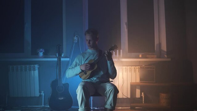 An adult smiling man plays the ukulele in the light neon dark room. A strong and blond guy sits and plays a small guitar in atmosphere place. A happy person plays a musical instrument.	